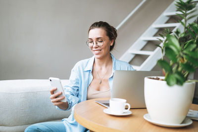 Smiling brunette woman with long hair working on laptop using mobile phone in bright home