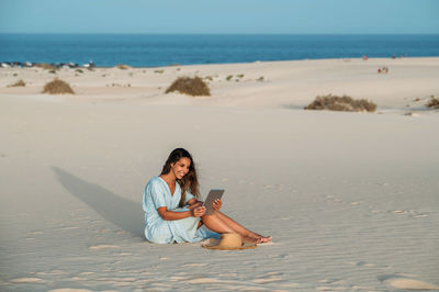 Sincere young barefoot female tourist with tablet sitting on sandy shore against sea and dunes of corralejo in sunlight in fuerteventura canary islands spain