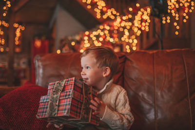 Candid authentic happy child in knitted beige sweater sitting with presents at lodge xmas decorated