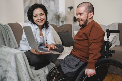 Smiling woman showing laptop to boyfriend in wheelchair at home