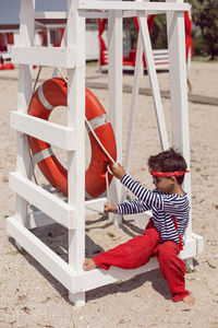 Child boy in striped clothes and red pants  standing on beach. white lifeguard tower, with a circle