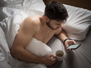 High angle view of man using phone while holding coffee cup on bed