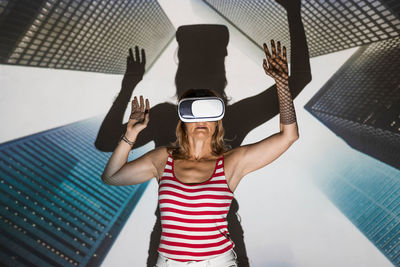 Unrecognizable female in casual wear and vr headset standing with arms raised against contemporary skyscrapers inside virtual city
