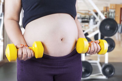 Midsection of pregnant woman holding yellow dumbbells in gym