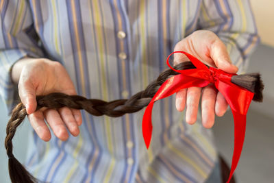 Long brown cut braid with red ribbon bow in female hands, donation of haircut, faceless concept