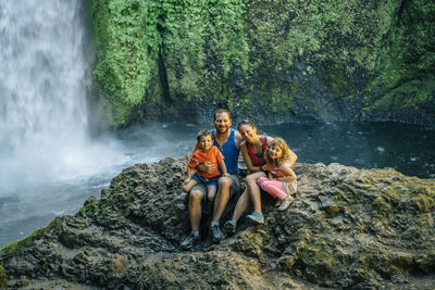 Portrait of happy family sitting on rocks against waterfall at forest