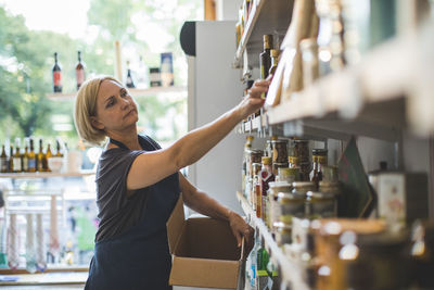 Mature female employee arranging bottles on shelf in deli