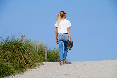 Rear view of woman walking on sand against clear blue sky at beach