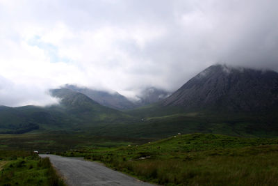 Country road leading towards mountains against cloudy sky