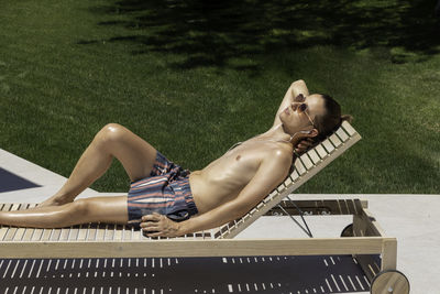 Young man on sunglasses and swimsuit lying on a sun lounger sun bathing close to the garden grass