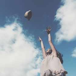 Low angle view of woman throwing hat in mid-air against cloudy sky