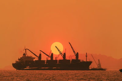 Silhouette cranes at harbor against orange sky