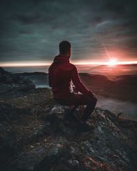 Rear view of man sitting on rock against sky during sunset