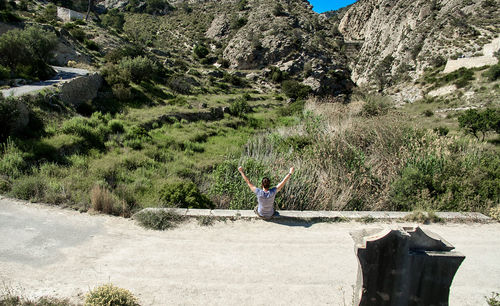 Rear view of young man sitting on rock against clear blue sky