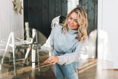 Young woman with blonde hair in grey sweater decorating christmas tree with gift box at house