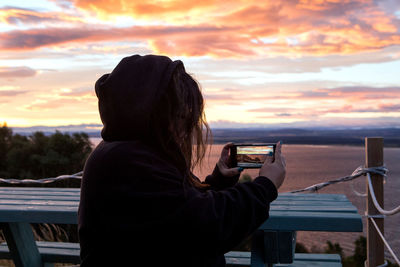 Woman photographing sea against sky during sunrise