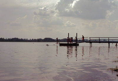 People on pier over lake against cloudy sky