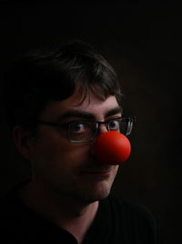 Portrait of man wearing eyeglasses with clown nose against black background