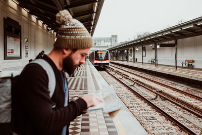 Side view of man wearing knit hat holding map standing at railroad station platform