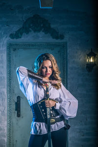 Woman in costume holding sword standing against door of abandoned building at night