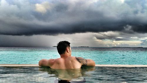 Rear view of man in infinity pool against cloudy sky