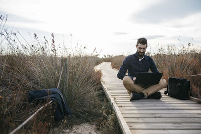 Full length of man using laptop while sitting amidst field on boardwalk