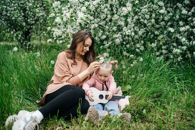 Young mother and baby daughter play the ukulele in a blooming apple orchard in nature