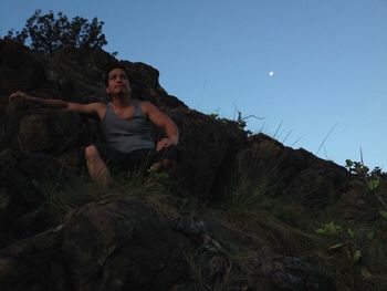 Low angle view of young man crouching on mountain against sky