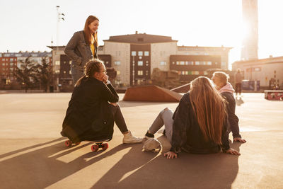 Teenage girl sitting on skateboard while talking with friends in park during sunny day