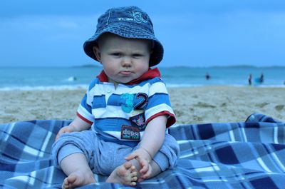 Portrait of baby boy sitting on blanket at beach against sky