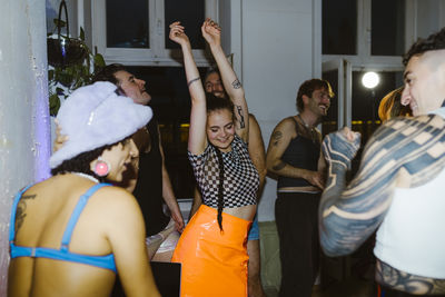 Smiling non-binary person dancing with arms raised amidst friends at nightclub