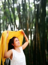 Woman holding sarong while looking up against trees in forest