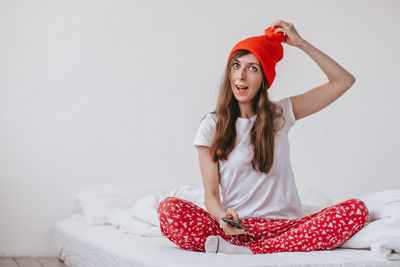 Smiling funny girl in a red hat and christmas pajamas sitting in bed with white sheets. 