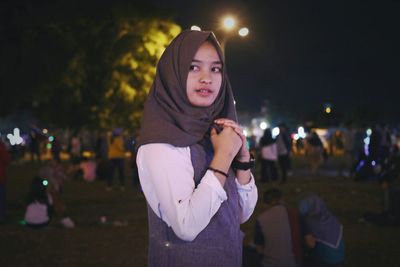 Portrait of young woman standing against illuminated tree at night