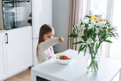 Pretty little girl with long hair sprinkles salt on vegetable salad in kitchen with bright interior 