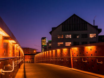 Illuminated street amidst buildings against sky at dusk