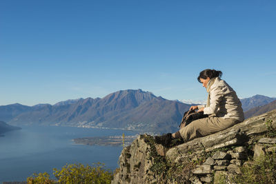 Side view of young woman sitting on rock against clear sky