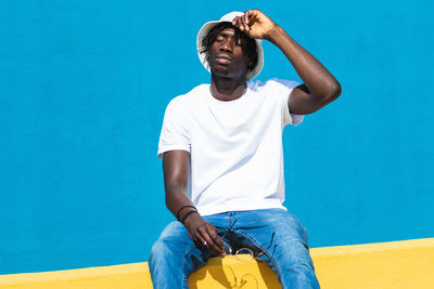 Young man looking away while sitting against blue wall