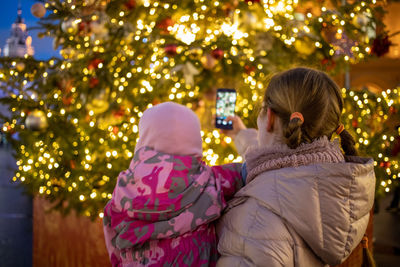 Mom and baby in her arms photographing a christmas tree at a street fair. back view