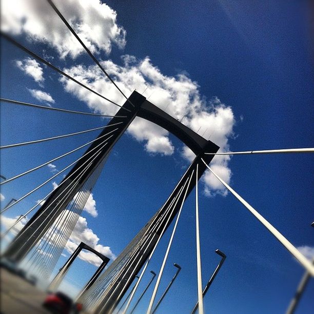 low angle view, sky, blue, built structure, architecture, cloud - sky, cloud, connection, day, building exterior, outdoors, transportation, part of, cloudy, no people, modern, engineering, bridge - man made structure, glass - material, cable