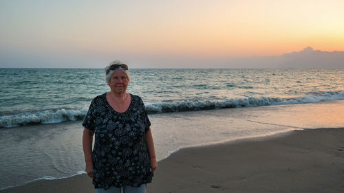 Portrait of senior woman standing at beach against sky during sunset