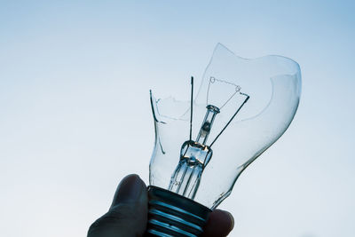 Close-up of hand holding broken light bulb against sky