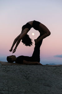 Side view of silhouette of flexible woman doing backbend and balancing on legs of man during acroyoga session against sunset sky with moon