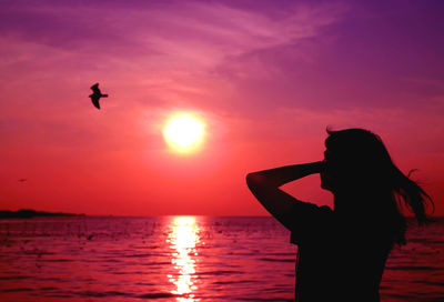 Silhouette of woman looking up to vivid purple pink colored sunrise sky with a flying bird