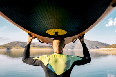Back view of unrecognizable mature male surfer standing with paddleboard above head near sea in evening and looking away