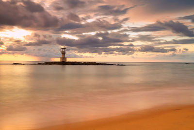 Lighthouse amidst sea under cloudy sky during sunset