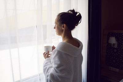 Woman in a white bathrobe is sitting on the edge of the bed and drinking tea from mug, large window 