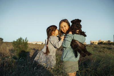 Two girl standing on field with dog against clear sky