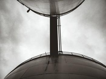 Low angle view of bridge connecting industrial tank against cloudy sky