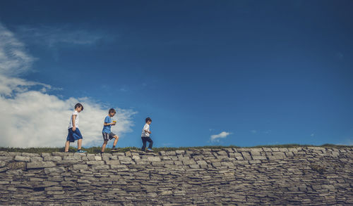 Low angle view of playful brothers walking on stone wall against sky